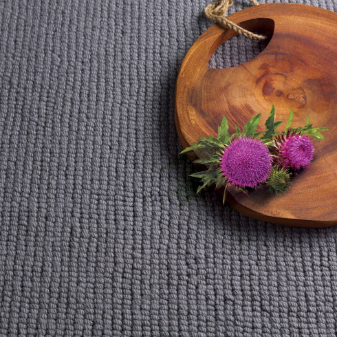 Close-up of Riviera Home Ben Nevis carpet in a rich grey tone, showcasing its chunky loop pile texture. Styled with a wooden board and Scottish thistle flowers for a natural, rustic touch.