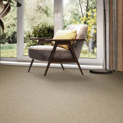 Luxury interior featuring Riviera Home Burford carpet in a warm natural tone. The textured wool loop pile flooring complements a modern wooden accent chair with a soft cushion, positioned by large glass doors overlooking a lush green garden.