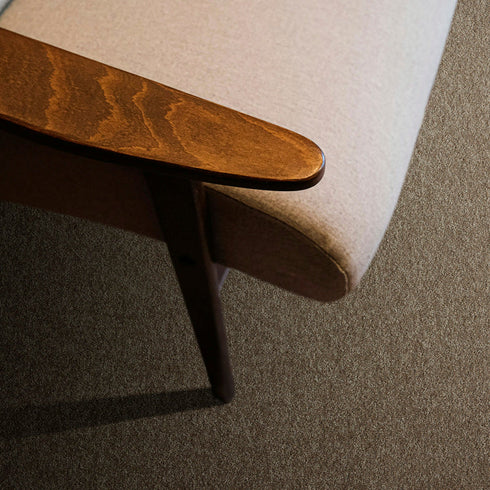 Lano Romance carpet in Cornstalk (a warm brown-beige), captured from a top-down perspective, showcasing its soft texture with a wooden armrest of a chair subtly visible in the frame, adding a natural and sophisticated touch.
