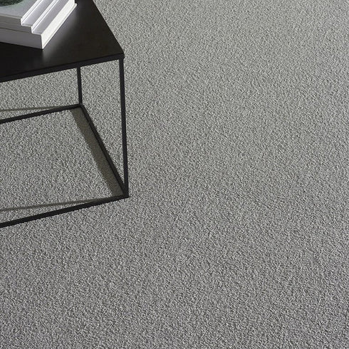 Lano Romance carpet in Silver (a soft grey), captured from a top-down view, showcasing its plush texture alongside a small table adorned with books and a candle, creating a cozy and elegant atmosphere.